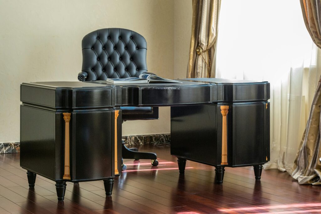 Black classic desk near elegant leather chair next to window with curtains in light study room